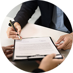 A person in a dark suit signing a document with a pen, with a hand visible from the opposite side.